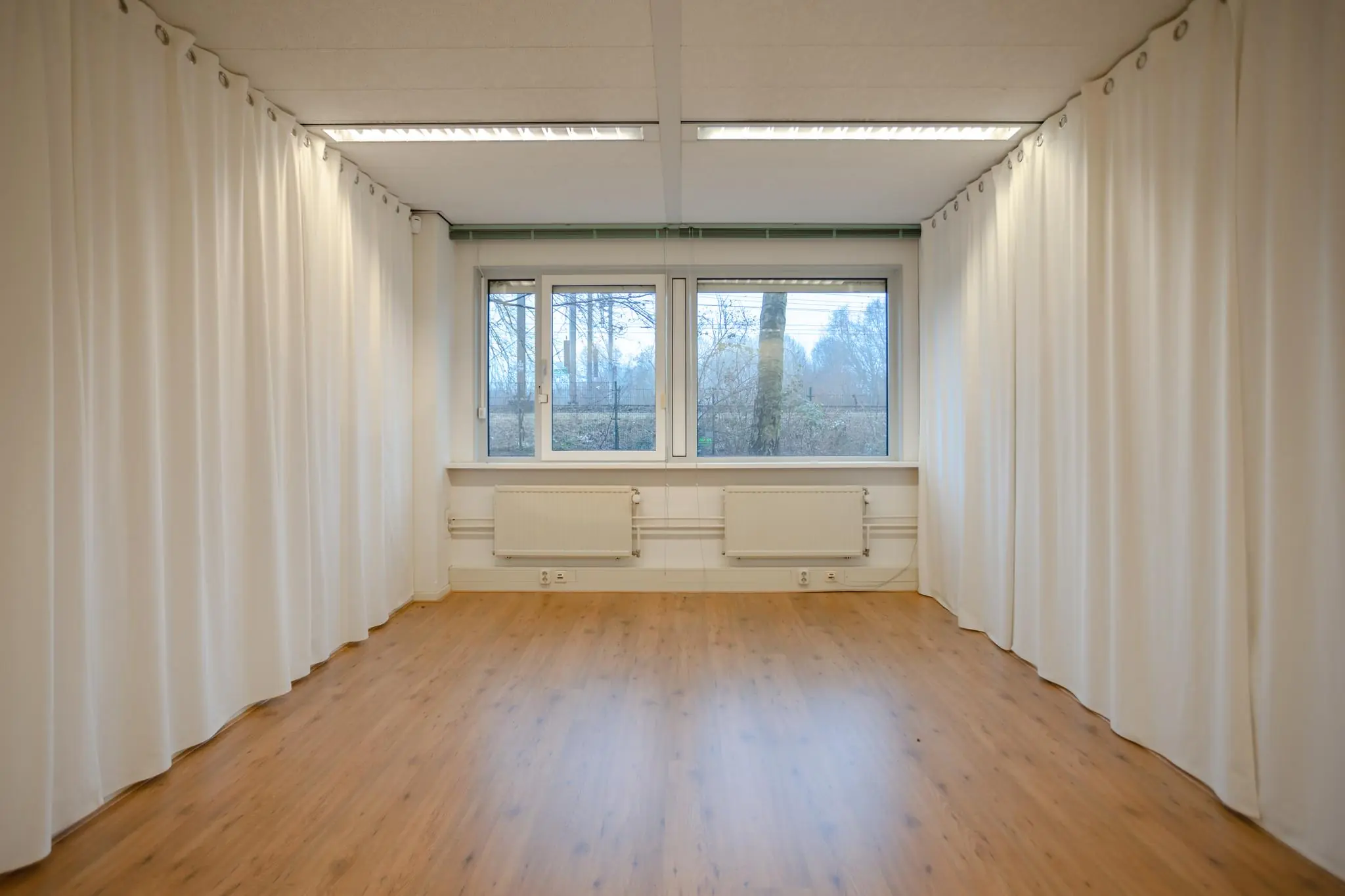 Lichte, lege ruimte met houten vloer, witte gordijnen en een groot raam aan de Christiaan Krammlaan.